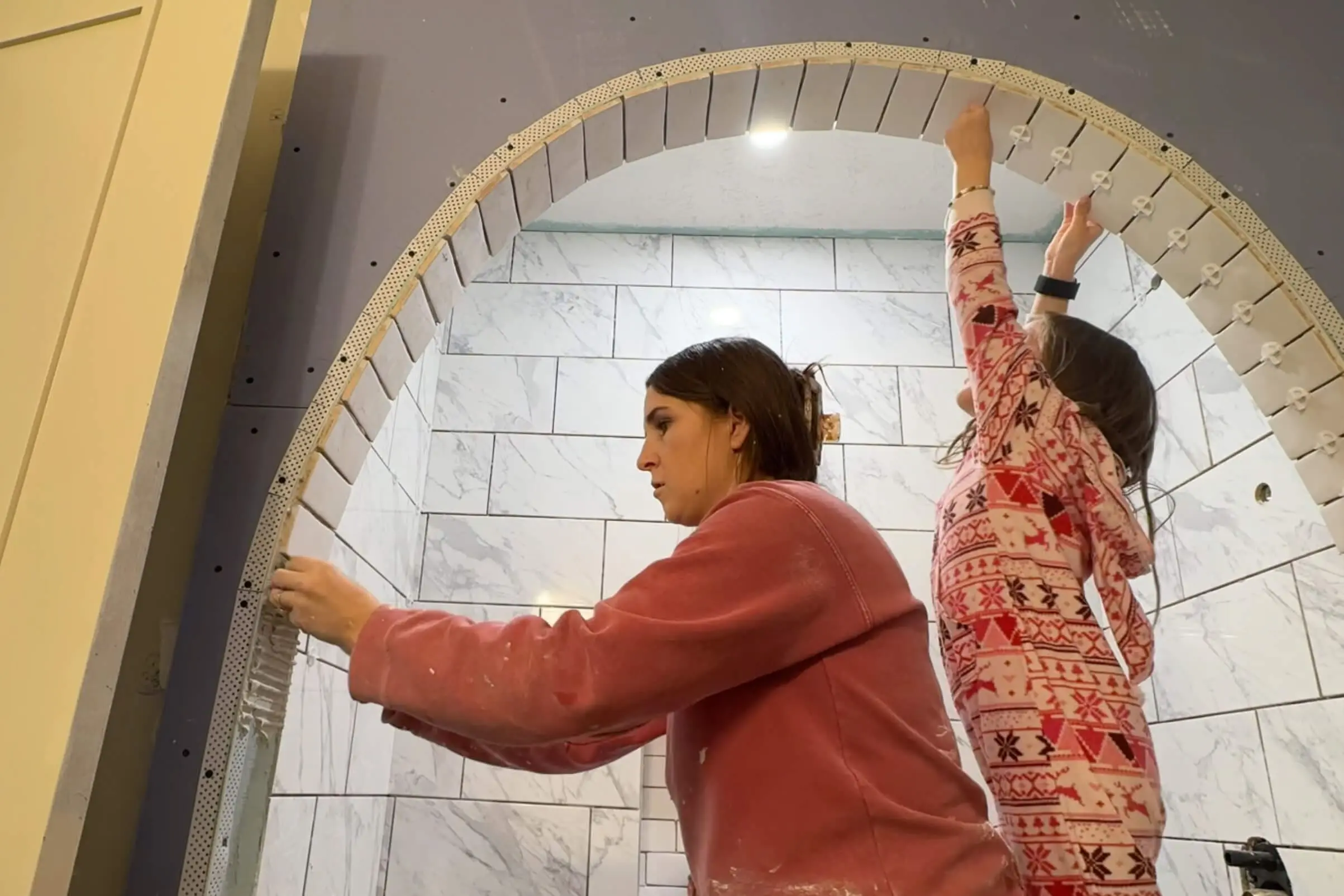 Two people working on tiling an arched bathroom shower. The adult is applying mortar to the curved edge, while a child in festive pajamas places a tile spacer at the top of the arch. The wall is tiled with large marble-style tiles, and the recessed niche is visible.